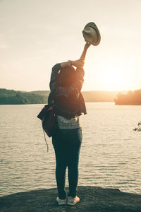 Rear view of woman holding hat at lakeshore against clear sky during sunset