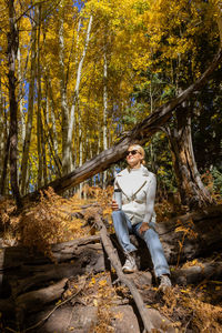 Pretty woman sits on fallen tree and drinks hot beverage from thermos travel mug in forest, nature