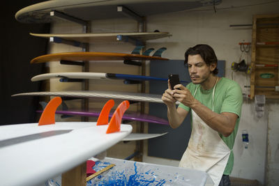 Male worker photographing surfboard through smart phone in workshop