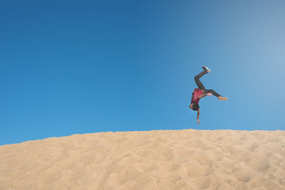 Full length of man jumping upside down on sand against clear blue sky