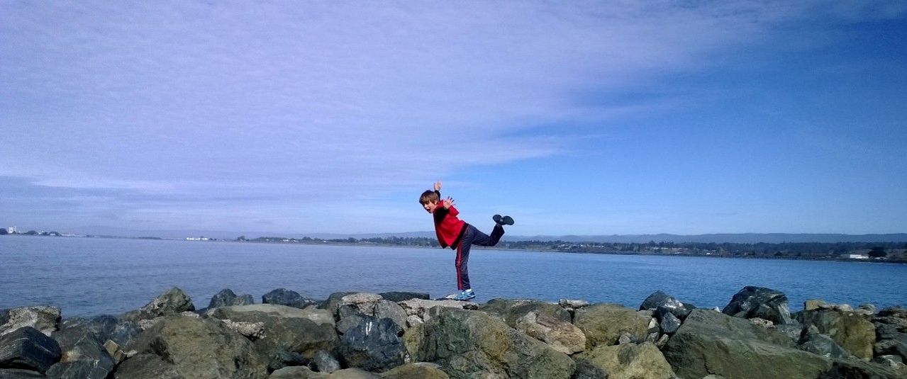 full length, water, lifestyles, leisure activity, sea, standing, casual clothing, rock - object, sky, rear view, scenics, tranquility, tranquil scene, beauty in nature, vacations, nature, young adult, arms outstretched