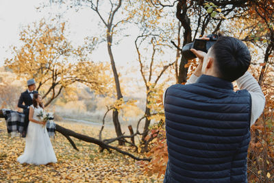 Rear view of woman photographing while standing against trees