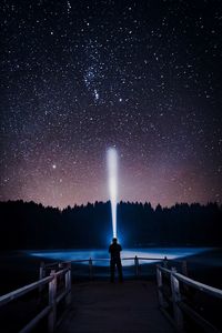 Rear view of man holding illuminated flashlight while standing against sky at night