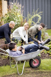 Boy using digital tablet while sitting in wheel barrow with family gardening in background