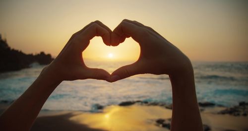 Cropped hand of woman making heart shape against sky during sunset