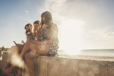 Female friends using phones by sea on retaining wall during sunny day