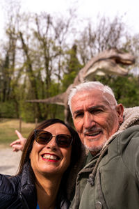 Happy middle aged couple taking a selfie in front a model t-rex in a jurassic park