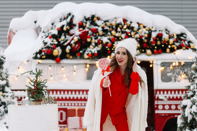 A beautiful woman with red lips in a warm hat stands by a decorated christmas van on the street