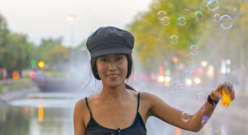 Portrait of smiling woman holding bubble wand while standing against pond