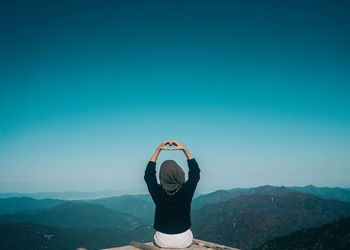 Rear view of woman making heart shape with hands while sitting on mountain 