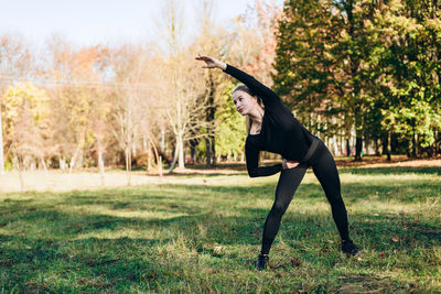 Girl in black sportswear performs exercise tilt left outdoors, autumn day.