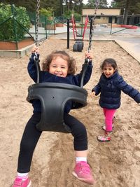 Portrait of happy girl enjoying on swing by sister standing at playground