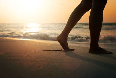 Low section of woman walking on beach against sky during sunset