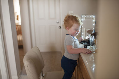 Side view of cute baby boy standing by beauty products on chair at home