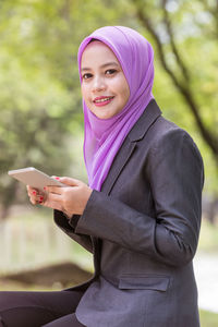 Portrait of young woman wearing hijab using mobile phone while sitting in park