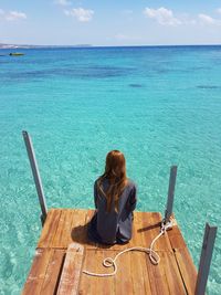 Rear view of woman sitting on jetty over sea against sky