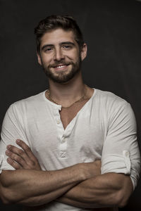 Portrait of smiling young man with arms crossed standing against black background