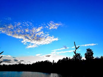 Silhouette trees by lake against blue sky