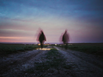 People walking on street amidst field against sky during sunset