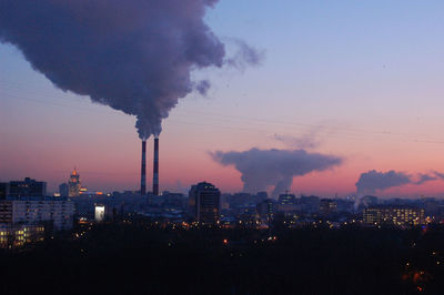 Smoke stack emitting in illuminated city against sky during sunset