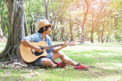 Young man playing guitar while sitting under tree on field at park during sunny day