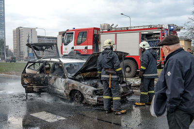 Firefighters standing by burnt car on road in city