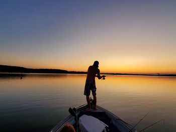 Silhouette fisherman fishing while standing on boat in lake against clear sky