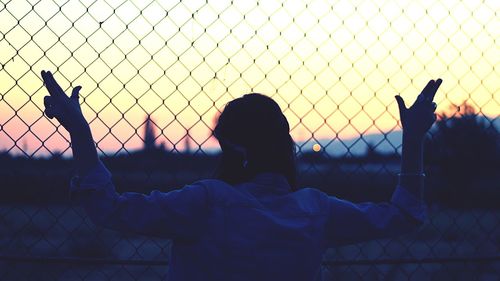 Rear view of man standing by chainlink fence against sky during sunset