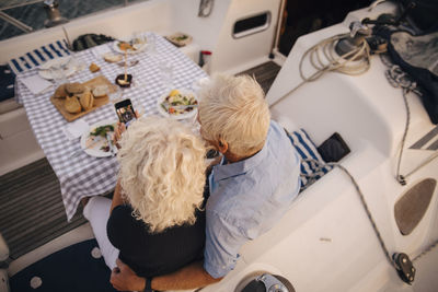 High angle view of senior couple taking selfie on mobile phone in sailboat during sunset