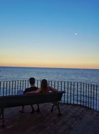 Rear view of couple sitting on bench by railing over sea against sky during sunset