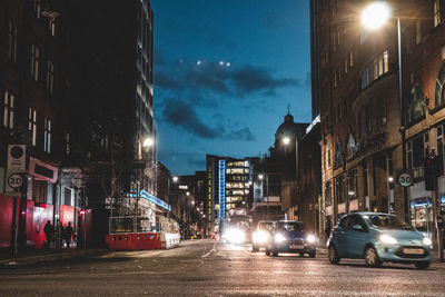 Illuminated city street by buildings against sky at night