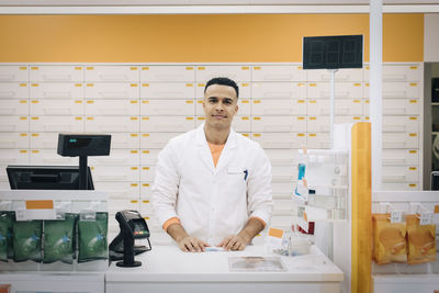 Portrait of confident male pharmacist standing at checkout at medical store