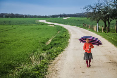 Full length of girl with umbrella walking amidst field on road