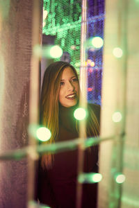 Smiling young woman standing by tree seen through illuminated lights