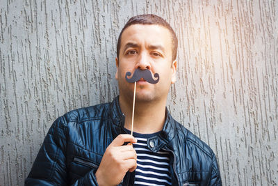 Portrait of man holding prop mustache against wall