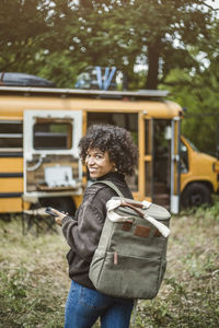 Rear view portrait of smiling young woman walking with backpack towards motor home in forest during camping