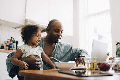 Father carrying daughter while using laptop during breakfast at table in dining room