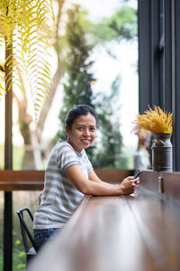Portrait of smiling woman sitting with mobile phone at table by window