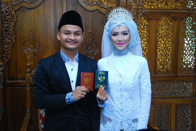 Portrait of bride and bridegroom holding passports while standing against wall