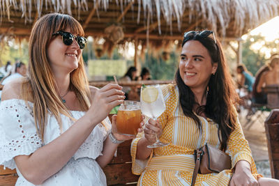 Two women sitting in beach bar at golden hour, having drinks and laughing