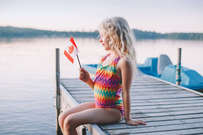 Girl sitting on pier by lake and waving canadian flag. kid celebrating canada day holiday outdoor