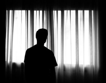 Rear view of silhouette man standing by curtain in darkroom at home