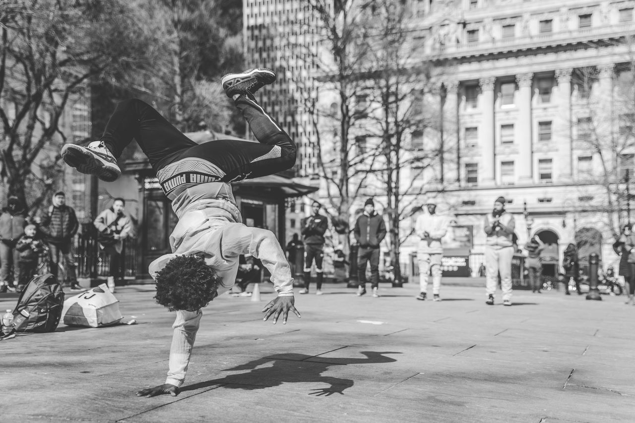 full length, real people, city, architecture, mid-air, men, leisure activity, lifestyles, day, skill, tree, jumping, focus on foreground, built structure, building exterior, people, nature, motion, incidental people, balance, outdoors, breakdancing, handstand