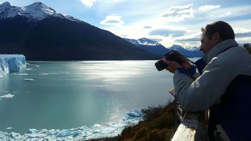Side view of man photographing by lake against sky