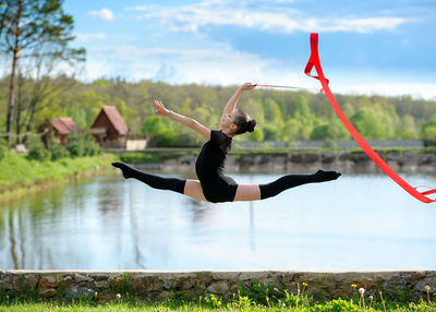 Side view of girl with ribbon jumping over retaining wall against lake