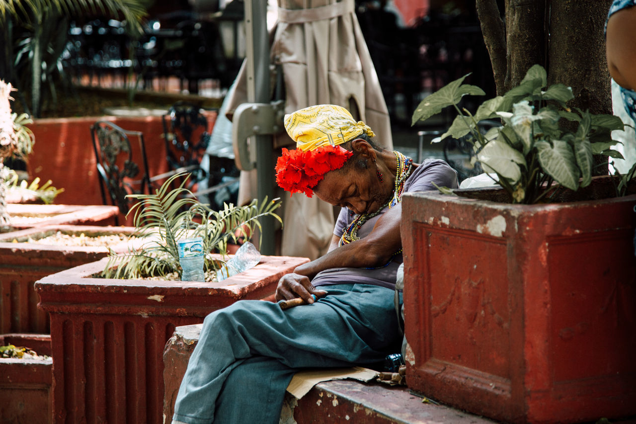 Havana, cuba - december 14, 2019: side view of poor aged female with flowers on head and cigar in hand sitting on bench next to street cafe and sleeping