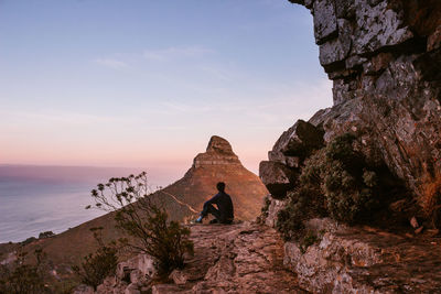 Young man sitting on rock against sky