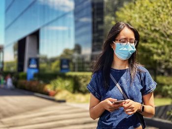 Portrait of asian woman in eyeglasses and surgical face mask using smartphone against building.