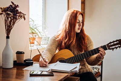 Unaltered candid portrait of young red haired woman in sweater playing acoustic guitar sitting by