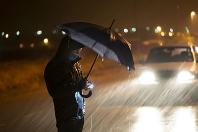 Young man with umbrella and face mask using mobile phone while standing against breakdown car during rainfall